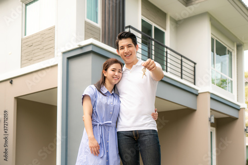 Portrait of Asian young couple standing and hugging together and holding house key looking happy in front of their new house to start new life. Family, age, home, real estate and people concept..