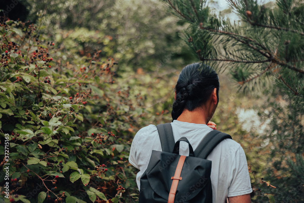 A young tanned man in a light grey T-Shirt, with long black hair and a backpack standing in a blackberry bramble bushes with his back to the camera A young tanned man in a light grey T-Shirt, with long black hair and a backpack standing in a blackberry bramble bushes with his back to the camera