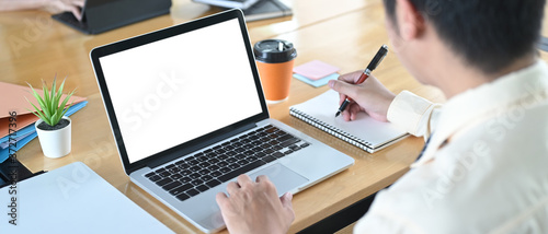 A man is using a white blank screen laptop while taking notes and sitting at the wooden table.
