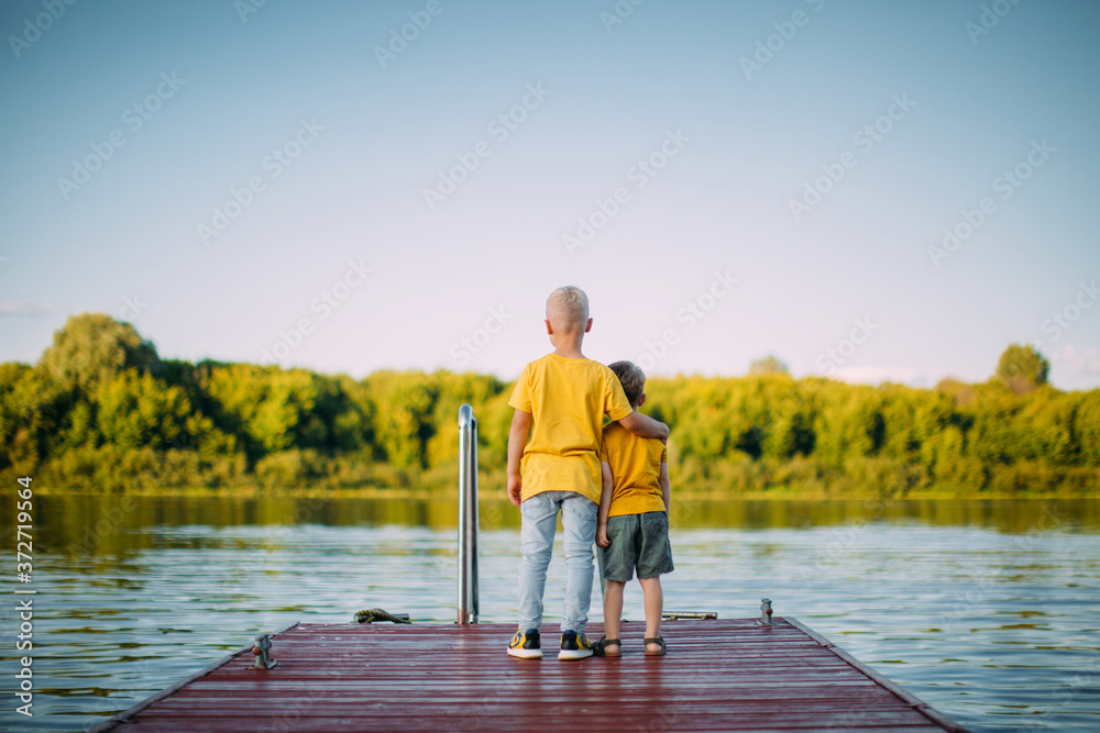 Cool kids brothers hug each other and look at the river staying on dock. Summertime photo about childhood