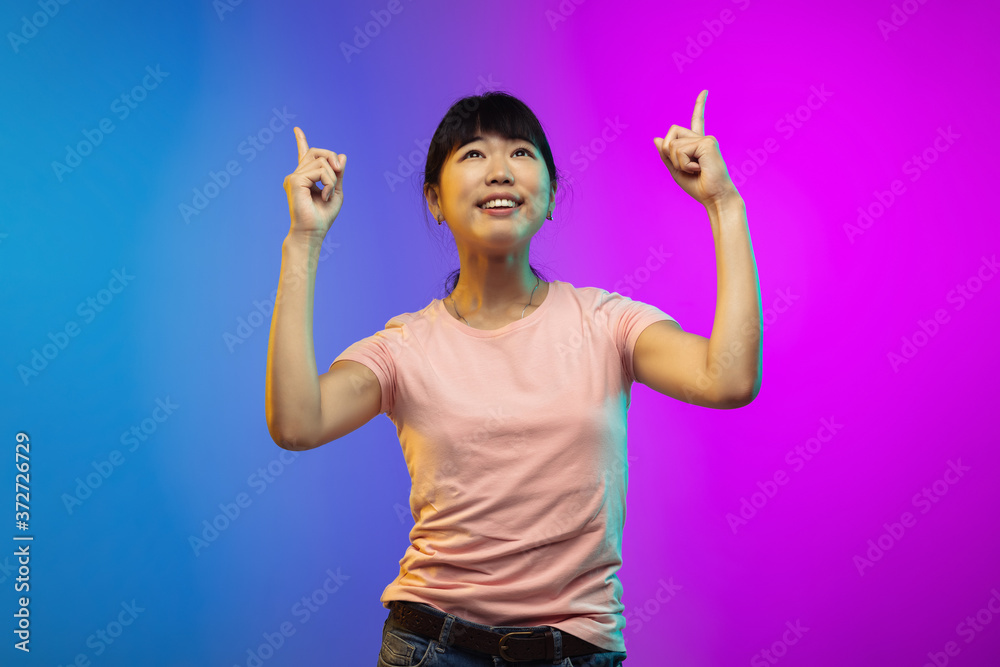 Pointing, showing. Asian young woman's portrait on gradient studio background in neon. Beautiful female model in casual style. Concept of human emotions, facial expression, youth, sales, ad. Pointing, showing. Asian young woman's portrait on gradient studio background in neon. Beautiful female model in casual style. Concept of human emotions, facial expression, youth, sales, ad.