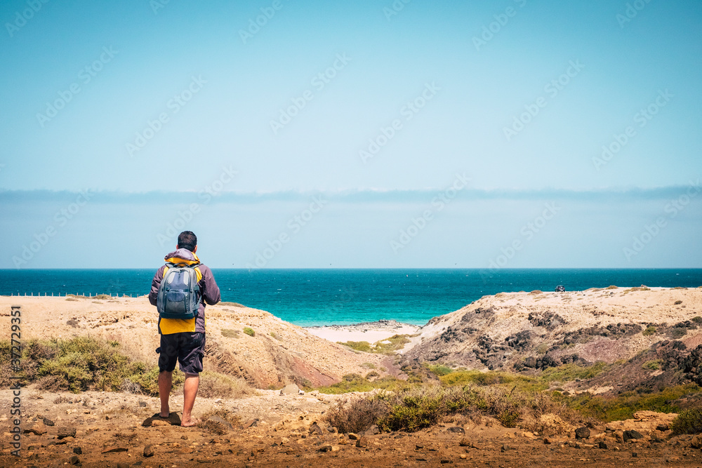 Back view of standing backpacker man looking at the beach and coast with blue sea and sky in background - concept of alternative explore lifestyle travel for active people