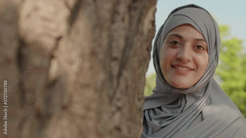 Tracking right of young Muslim woman with no makeup wearing hijab is standing near tree in park, looking at camera, waving her hand and smiling