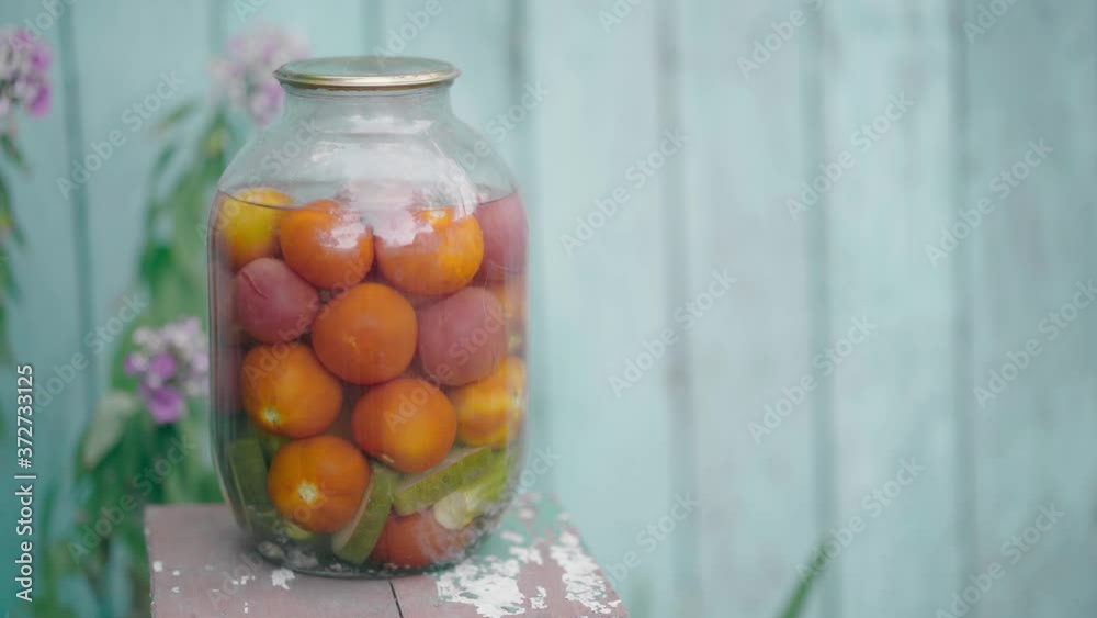 Big glass jar with pickled vegetables on chair on background of old wooden house. Canned tomatoes and cucumbers in sealed jar on street in garden. Big glass jar with pickled vegetables on chair on background of old wooden house. Canned tomatoes and cucumbers in sealed jar on street in garden.