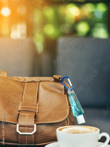 Mini alcohol gel bottle to kill Corona Virus(Covid-19) hang on a brown leather shoulder bag with hot coffee on table in cafe.New normal lifestyle.Health care concept.Selective focus on alcohol gel