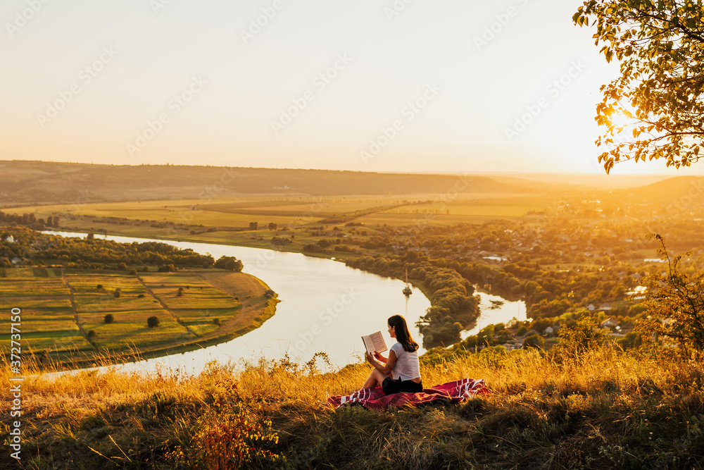 Young girl sitting on a hill with beautiful landscape and reading book. Sunset. Romantic evening. Loneliness.