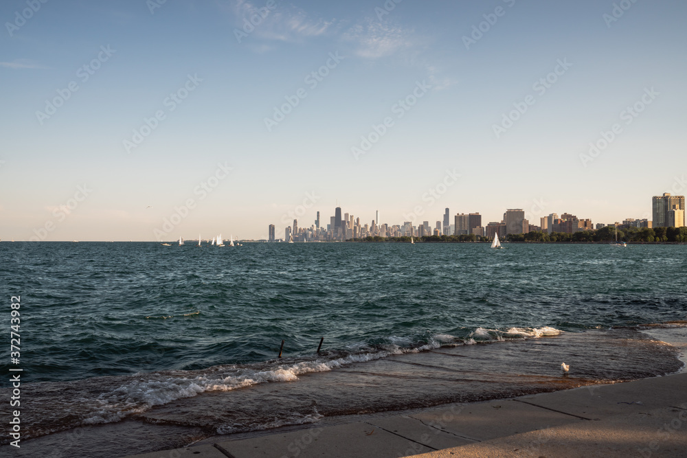 Sailboats and boats float across the Lake Michigan water in front of the magnificent Chicago skyline with prominent and historic high rise buildings and architecture on a sunny summer day with waves.