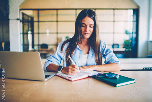 Pensive female student learning and writing information in notepad during online courses using laptop, skilled freelancer making notes of ideas updating application on netbook sitting in office