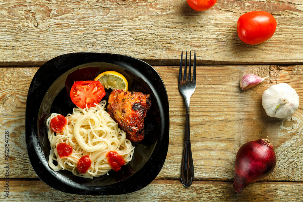 Black plate of spaghetti with baked chicken with a fork on a wooden table near vegetables. Black plate of spaghetti with baked chicken with a fork on a wooden table near vegetables.