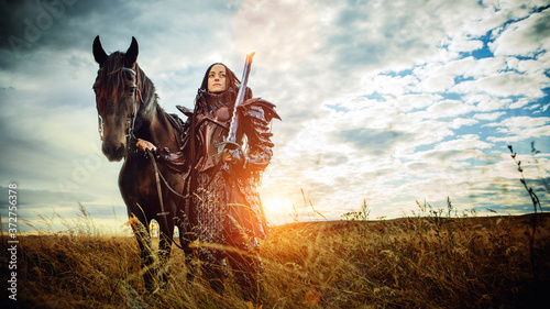 Fotografie Girl in medieval knight's armor with a horse against the sunset fields backgroun