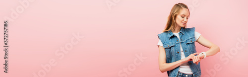 panoramic shot of young woman in denim clothes touching smartwatch isolated on pink