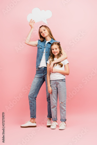 full length view of mother in denim clothes holding thought bubble while embracing child in white t-shirt and jeans on blue