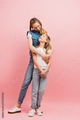 full length view of young woman in denim clothes embracing daughter while looking at each other on pink