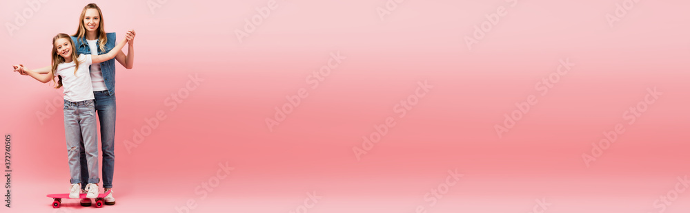 horizontal image of mother supporting daughter standing on penny board by holding her hands on pink horizontal image of mother supporting daughter standing on penny board by holding her hands on pink