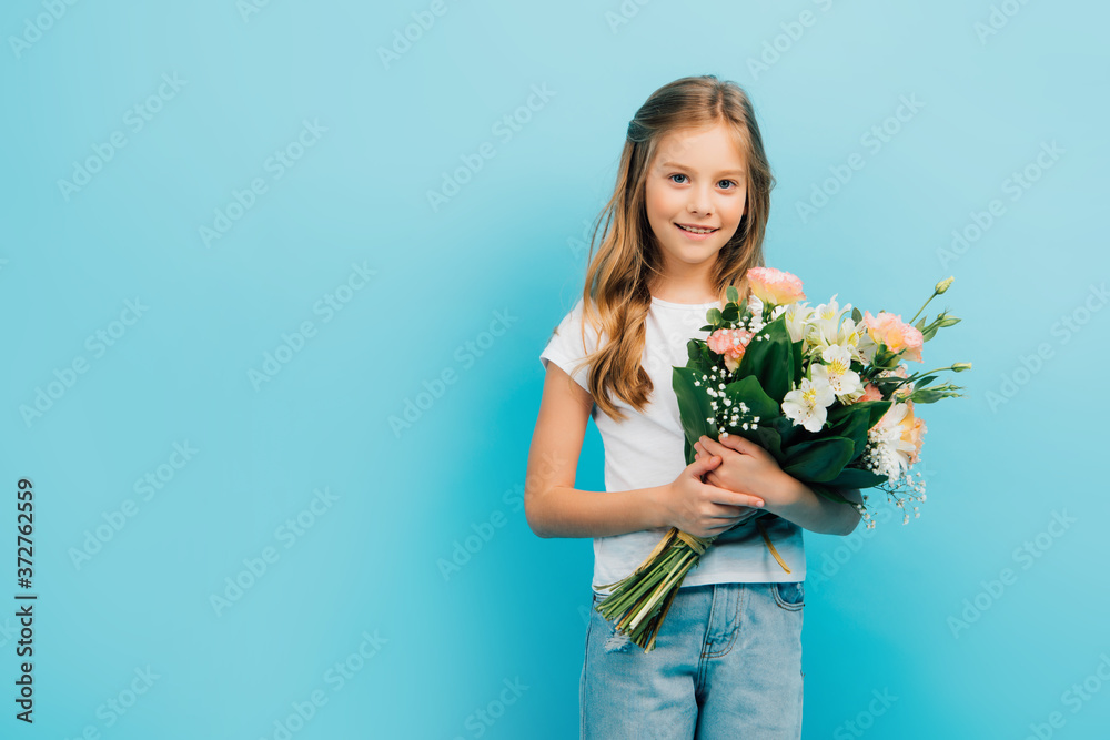 child in white t-shirt and blue jeans looking at camera while holding bouquet isolated on blue child in white t-shirt and blue jeans looking at camera while holding bouquet isolated on blue