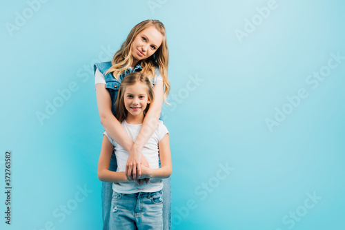 mother embracing child wearing white t-shirt and jeans while looking at camera isolated on blue