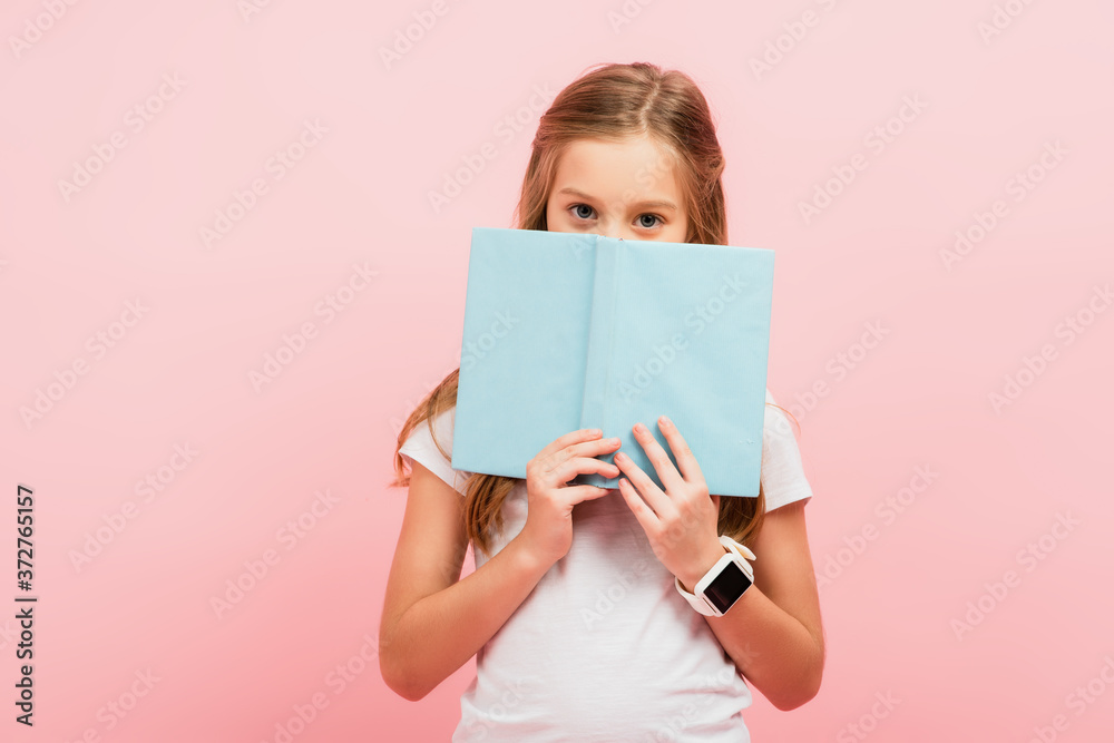 child in smartwatch obscuring face with book while looking at camera isolated on pink child in smartwatch obscuring face with book while looking at camera isolated on pink