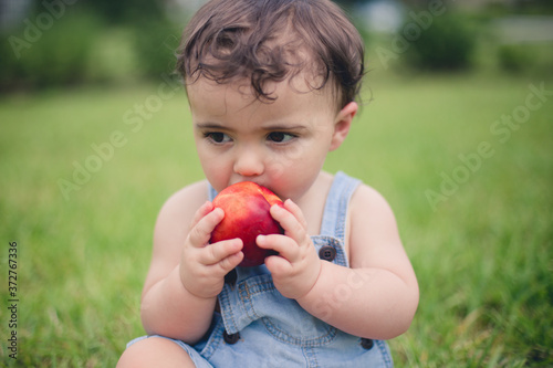 Baby eating a apple or peach outside in the backyard of his home in the grass wearing his overalls 