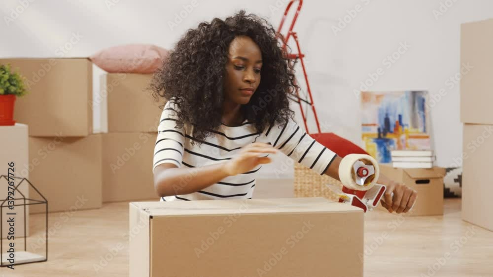 Beautiful young African-American woman is intently preparing to movein new apartment. Confident girl closes cardboard box with scotch tape. Beautiful young African-American woman is intently preparing to movein new apartment. Confident girl closes cardboard box with scotch tape.