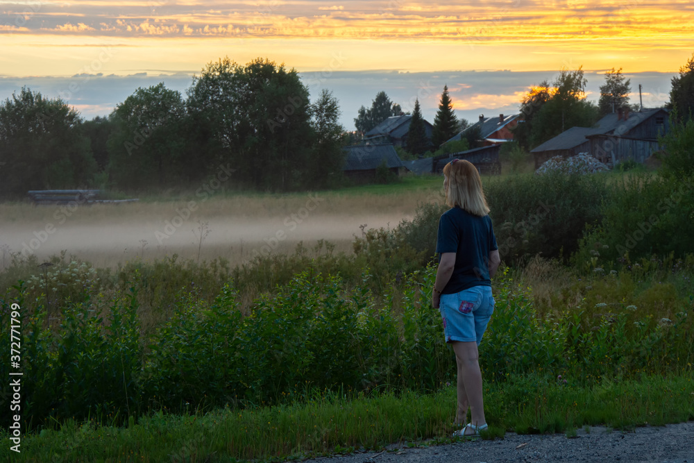 A young red-haired woman on a summer evening looks at the rural landscape. Fog over a field in the village. Shallow depth of field A young red-haired woman on a summer evening looks at the rural landscape. Fog over a field in the village. Shallow depth of field