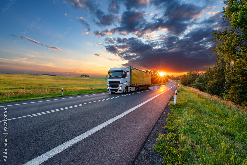 Truck driving on the asphalt road in rural landscape in the rays of the sunset with dark storm cloud