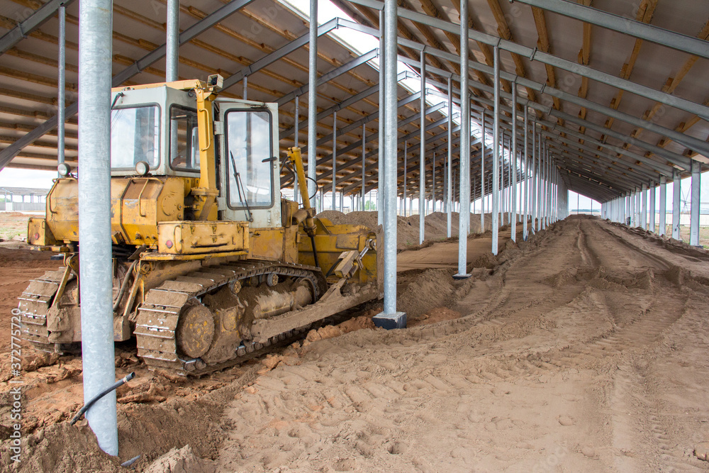 Fototapeta premium A bulldozer is leveling the ground in a barn under construction. Construction of an agricultural complex. Earthworks using special equipment. Bulldozer is pushing soil in a worksite