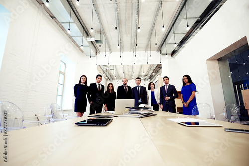 Young creative multi ethnic group of architects posing while standing near table with open laptop computer and paper documents, purposeful business men and women in formal wear resting after briefing