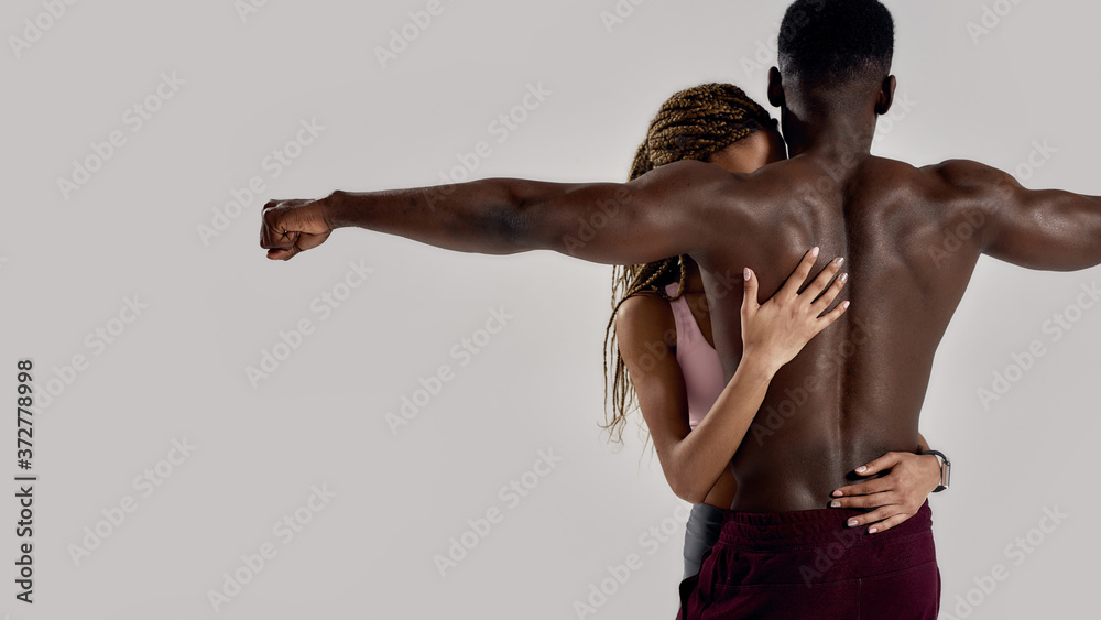 Sportive mixed race woman embracing, touching muscular african american man back while posing together isolated over grey background. Sports, workout, bodybuilding concept