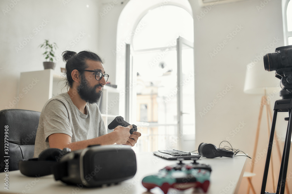 Male technology blogger in glasses looking dissatisfied, holding game controller joystick while recording video blog or vlog about new gadgets at home studio
