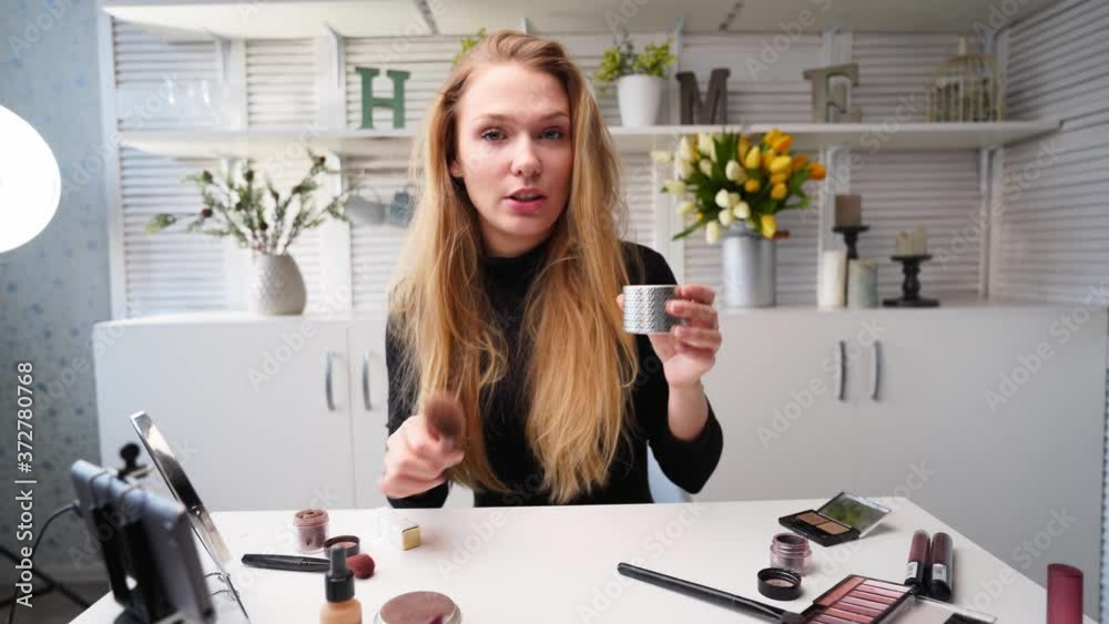 Beauty blogger woman filming daily makeup routine tutorial at camera on tripod. Influencer lady live streaming cosmetics product review in home studio. Vlogger female applies skin powder with brush.
