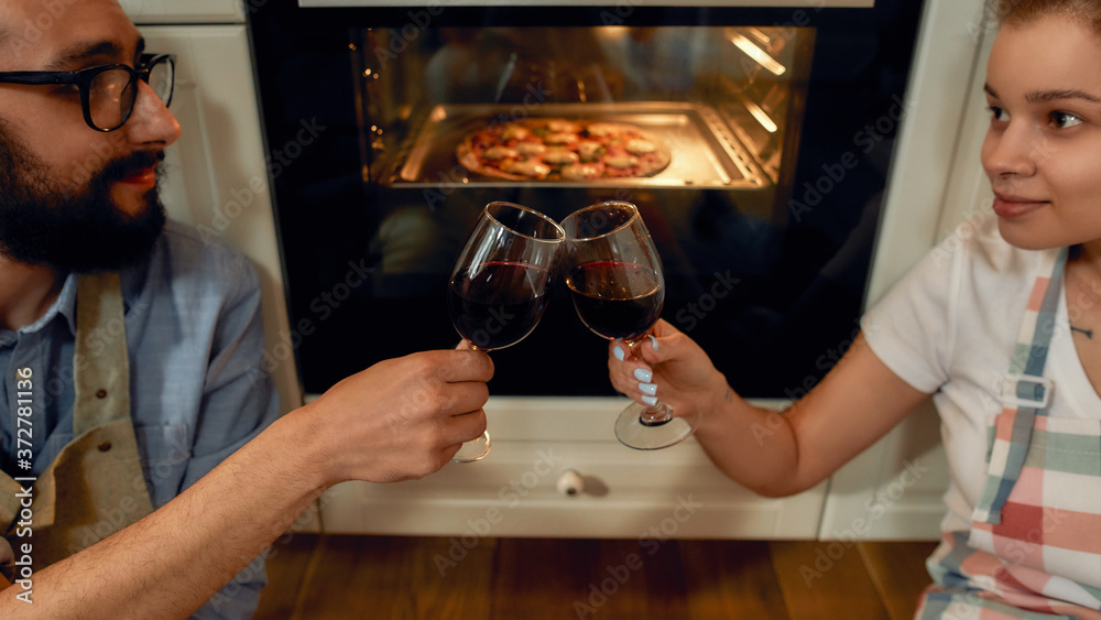 Close up shot of young happy couple having glass of wine while cooking in the kitchen. Man and woman in apron looking at each other, waiting for pizza to be baked