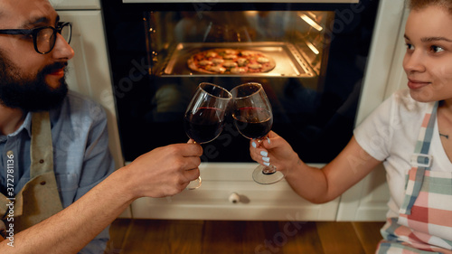 Close up shot of young happy couple having glass of wine while cooking in the kitchen. Man and woman in apron looking at each other  waiting for pizza to be baked