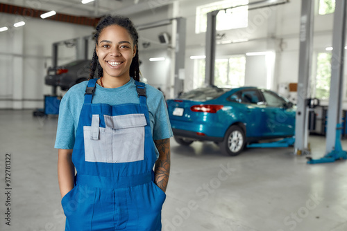 Best quick help. Portrait of young african american woman, professional female mechanic in uniform smiling at camera, standing in auto repair shop. Car service, repair, maintenance and people concept