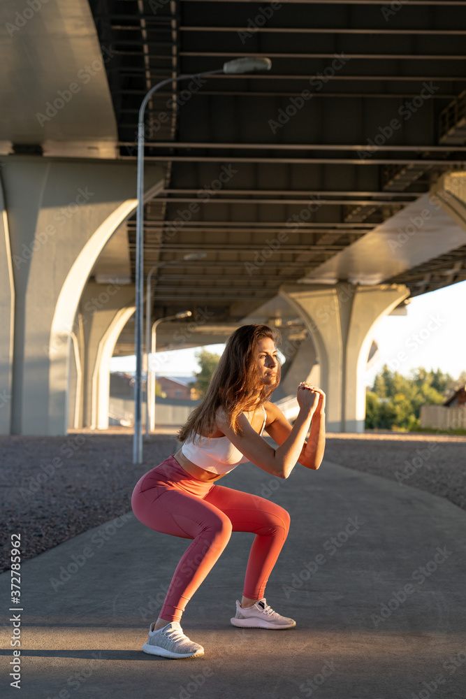 Young adult sporty woman in pink leggings squatting, making cardio workout, has exercises outdoors, under the bridge at sunset. Deep squat. 