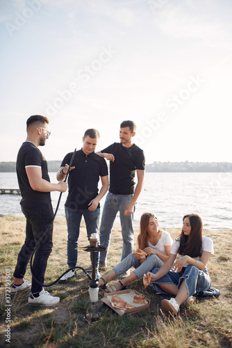 Group of friends is sitting near the lake. They wear casual clothes. Young people eat pizza outside.