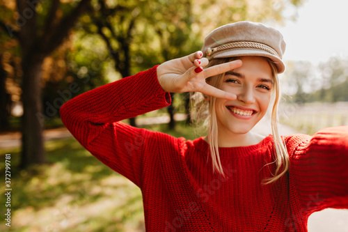 Pretty blonde girl in stylish hat and red sweater posing with smile to make a selfie in the park.