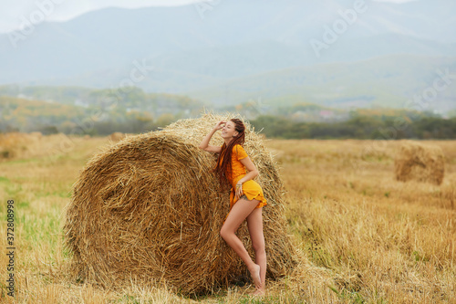 Redhead woman in short orange dress. Cute smiling russian woman posing in a field near a haystack. Girl in sensual short orange dress