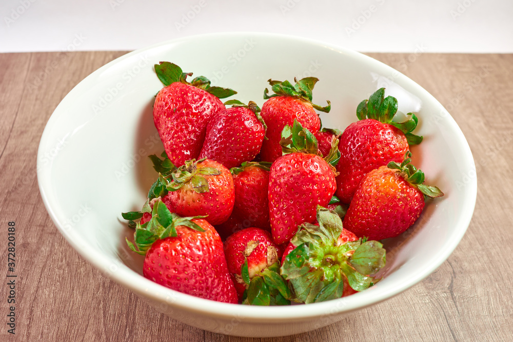 Red and juicy strawberries in a white bowl on a wooden board and white background Red and juicy strawberries in a white bowl on a wooden board and white background