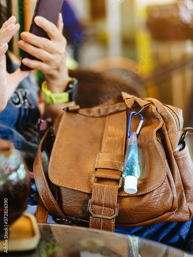 Mini portable alcohol gel bottle to kill Corona Virus(Covid-19) hang on a brown leather shoulder bag with hot coffee in cafe.New normal lifestyle.Health care concept.Selective focus on alcohol gel
