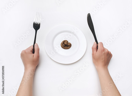 Women's hands hold a stainless knife and fork near plate with artificial feces. Fake news, jokes. The concept of inept cook, crappy restaurant, bad food, shit taste. © shchus