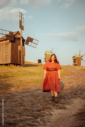 Beautiful and confident plus size model in red dress and brown boots posing outdoors at autumn landscape with wooden mill at background