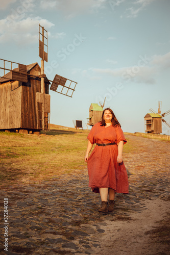 Beautiful and confident plus size model in red dress and brown boots posing outdoors at autumn landscape with wooden mill at background