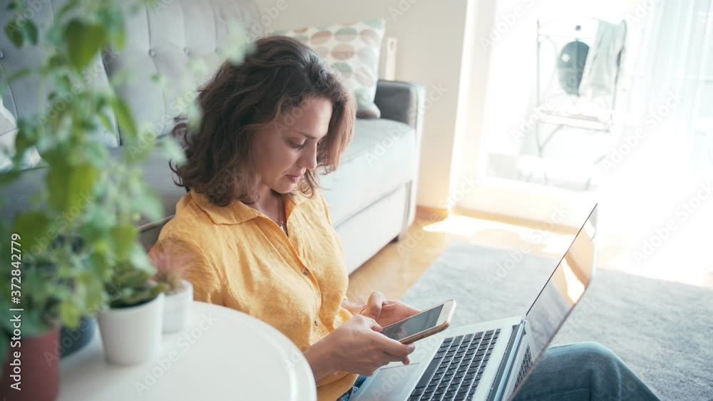 Young cheerful woman freelancer working from home. Woman typing a message on her phone while sitting on the floor near the couch with a notebook on her laps. Young cheerful woman freelancer working from home. Woman typing a message on her phone while sitting on the floor near the couch with a notebook on her laps.