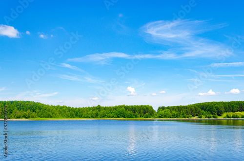 Lake in the forest on a clear summer day. Specular reflection in the water. Green foliage of trees. Coast. Blue sky with white clouds on a sunny day.