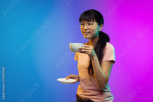 Coffee time. Asian young woman's portrait isolated on gradient studio background in neon. Beautiful female model in casual style. Concept of human emotions, facial expression, youth, sales, ad.