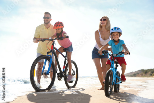 Happy parents teaching children to ride bicycles on sandy beach near sea