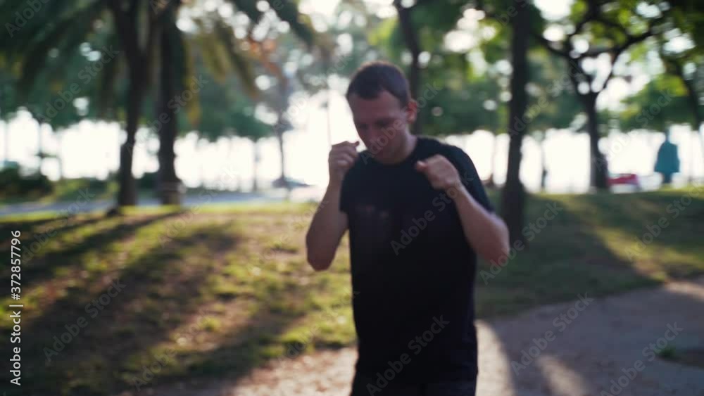 Morning workout. Muay thai box training in the park. Young man is running and practicing punches in the sunny urban park. Sport activity. Athlete. Punch, elbow, knee. Closeup of punch practice. Morning workout. Muay thai box training in the park. Young man is running and practicing punches in the sunny urban park. Sport activity. Athlete. Punch, elbow, knee. Closeup of punch practice.
