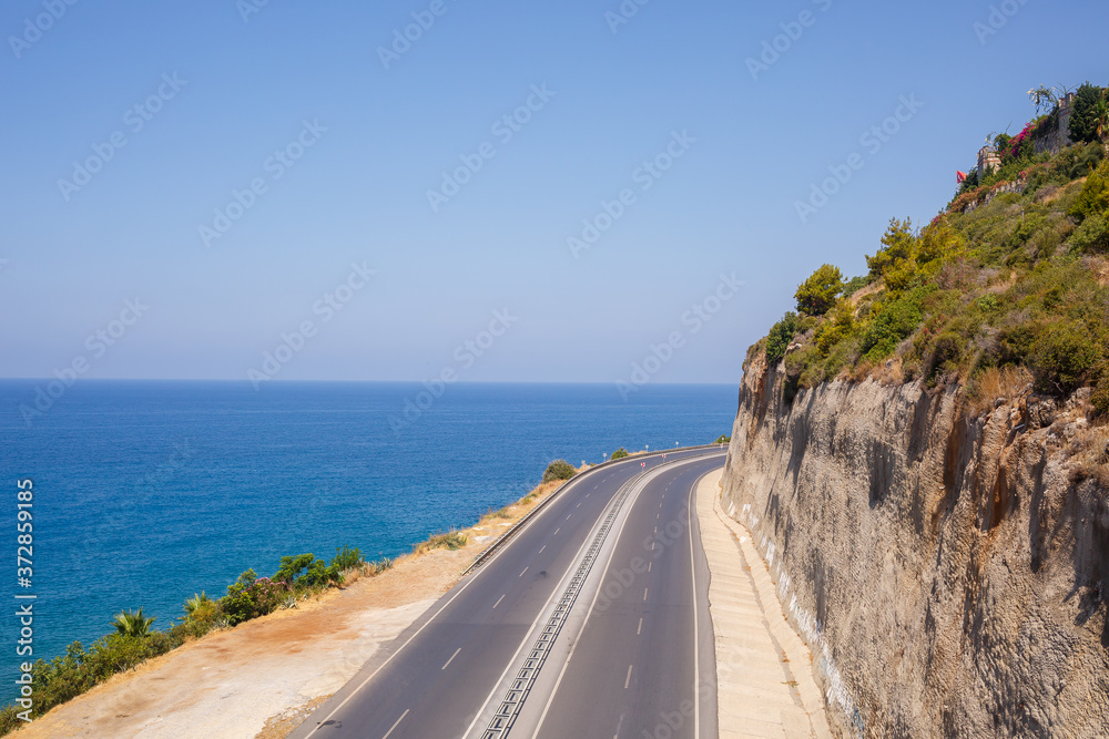 An empty asphalt road winds along the breathtaking scenic coastline on a sunny summer day. A spectacular shot of a coastal road overlooking clear blue skies and the calm Mediterranean Sea.