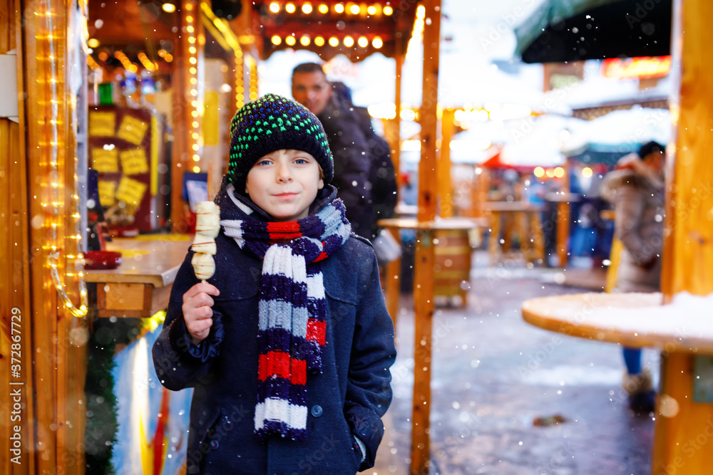 Little cute kid boy eating white chocolate covered fruits on skewer on traditional German Christmas market. Happy child on traditional family market in Germany during snowy day.