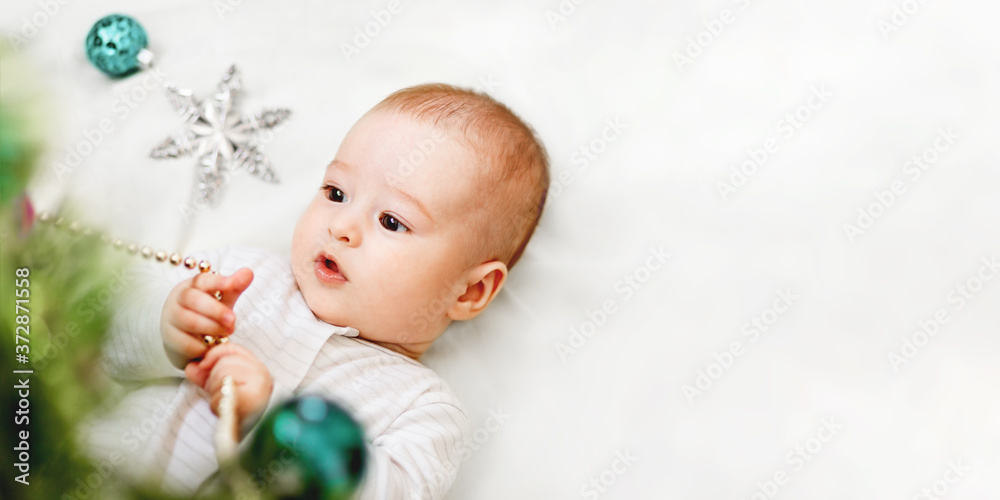 Baby boy or girl lying under Christmas tree. Little child plays with fir tree decorated for New Year celebration. Cute kid portrait. Winter holiday spirit. Banner with copy space. Baby boy or girl lying under Christmas tree. Little child plays with fir tree decorated for New Year celebration. Cute kid portrait. Winter holiday spirit. Banner with copy space.